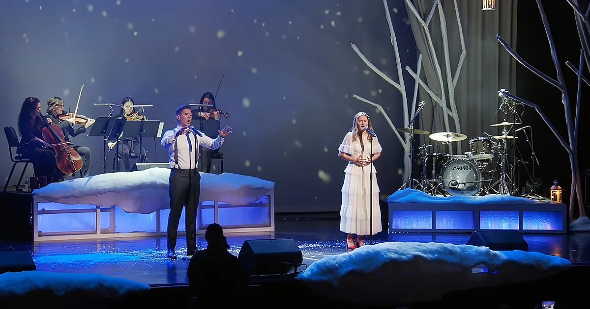 A man and woman sing on stage with a snowy backdrop and musicians playing.