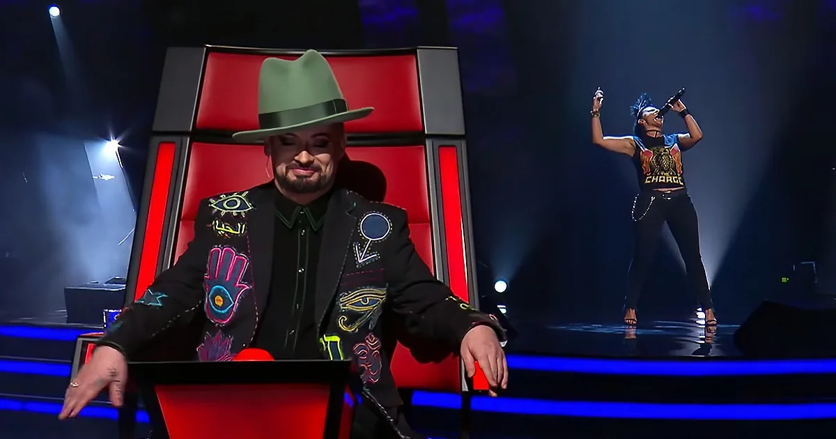 A man in a green hat and colorful jacket sits on a red chair, watching a performer on stage.