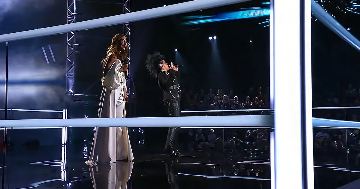 A woman in a white dress stands on stage, smiling, while another woman in black sings into a microphone.