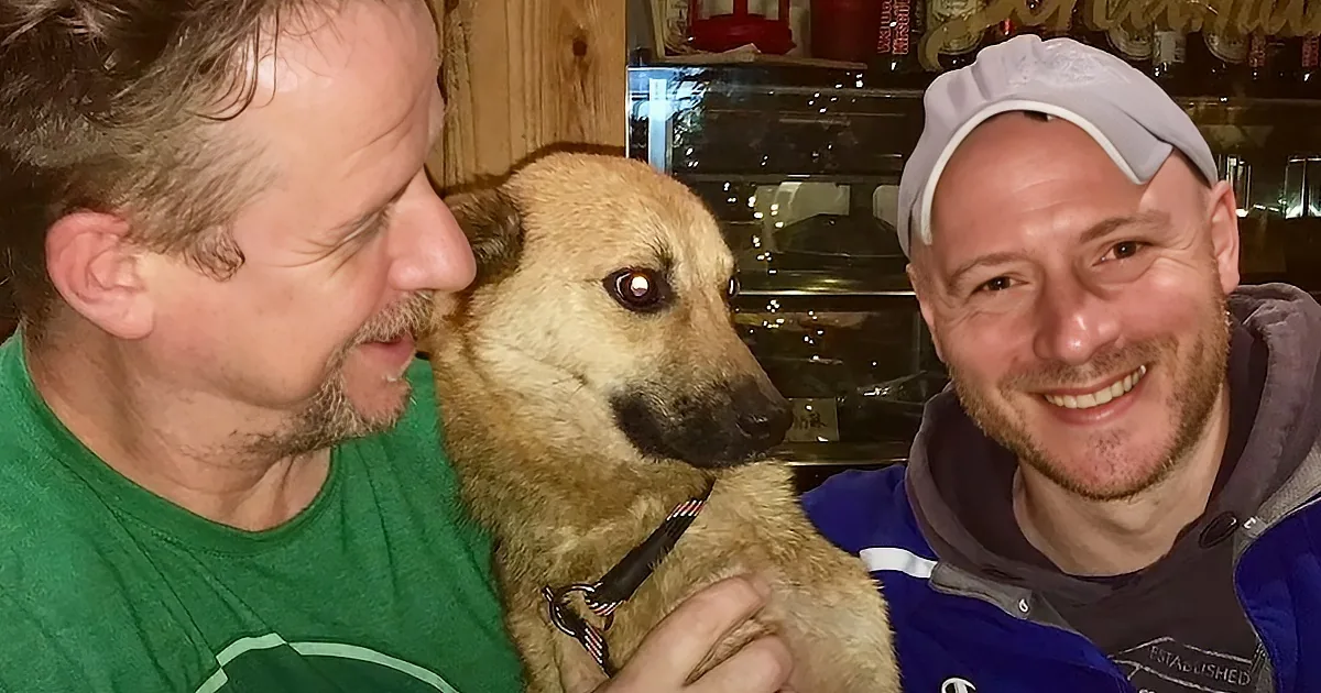 Two men smile with a dog, one holding it, in a cozy indoor setting.