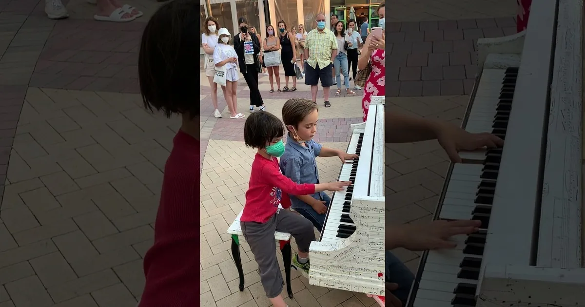 Two children play a piano together, one wearing a mask, in a public setting.