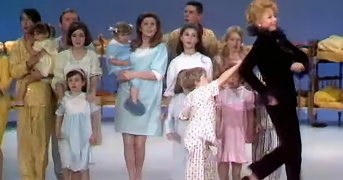 A woman in black directs a group of people in colorful attire, some holding children, in a staged scene.