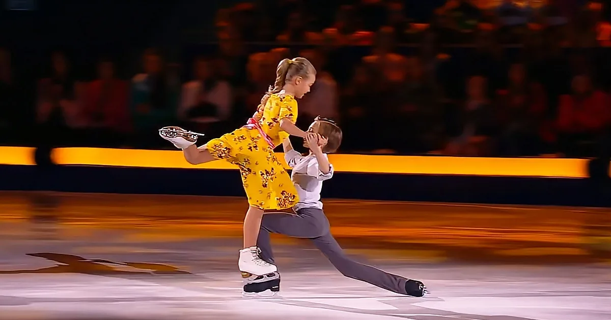 A figure skater lifts a partner in a dynamic pose on ice, both wearing colorful costumes.