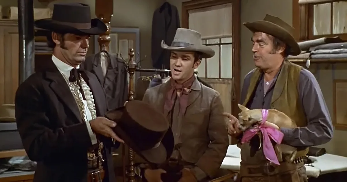 Three men in cowboy hats and western attire are gathered, one holding a hat, another a dog with a pink ribbon.