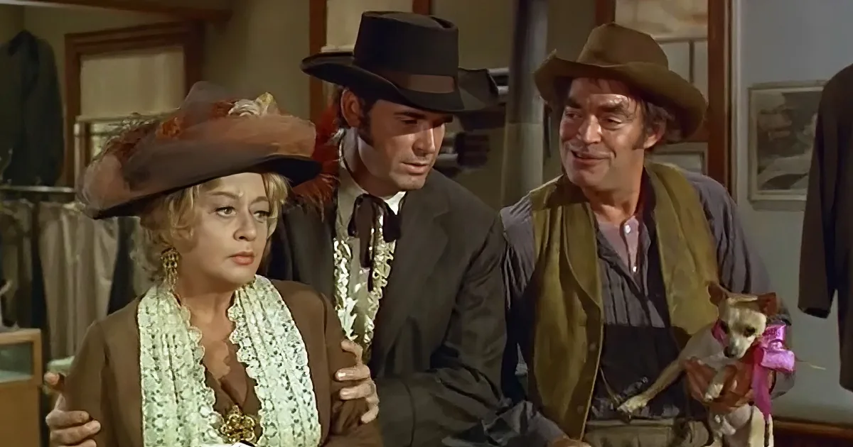 A woman in a brown coat and hat is being embraced by two men in cowboy attire, one holding a small dog.
