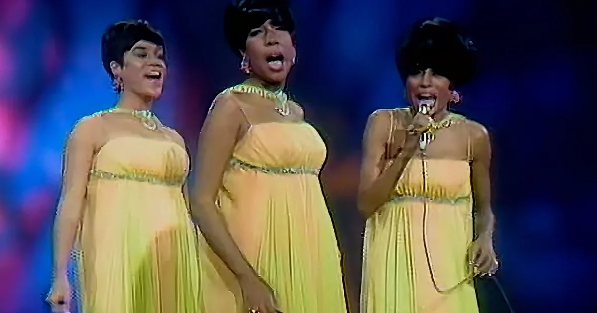 Three women in yellow dresses sing on stage, one holding a microphone.