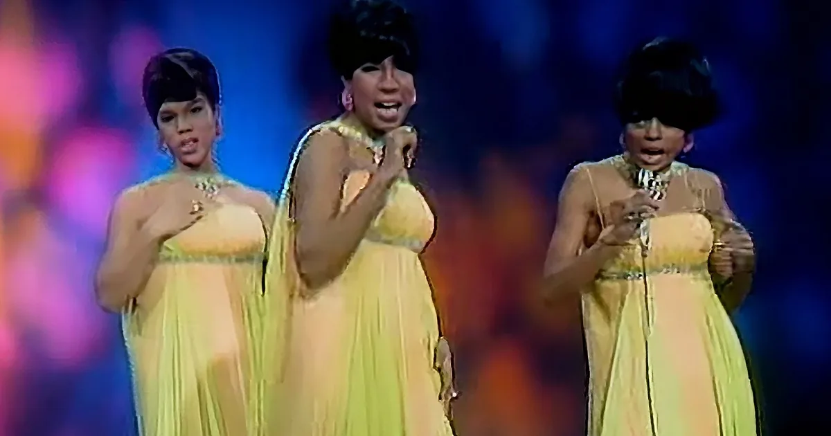 Three women in yellow dresses sing on stage, holding microphones, with colorful lights in the background.