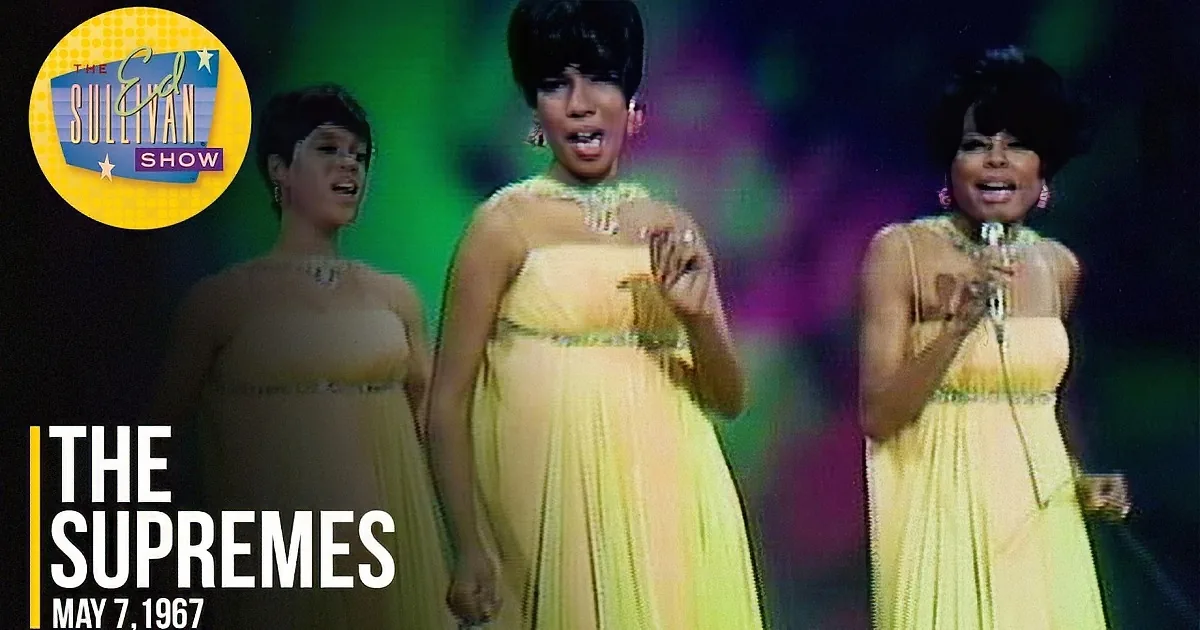 A woman in a yellow dress sings into a microphone, wearing a sparkling necklace and earrings.