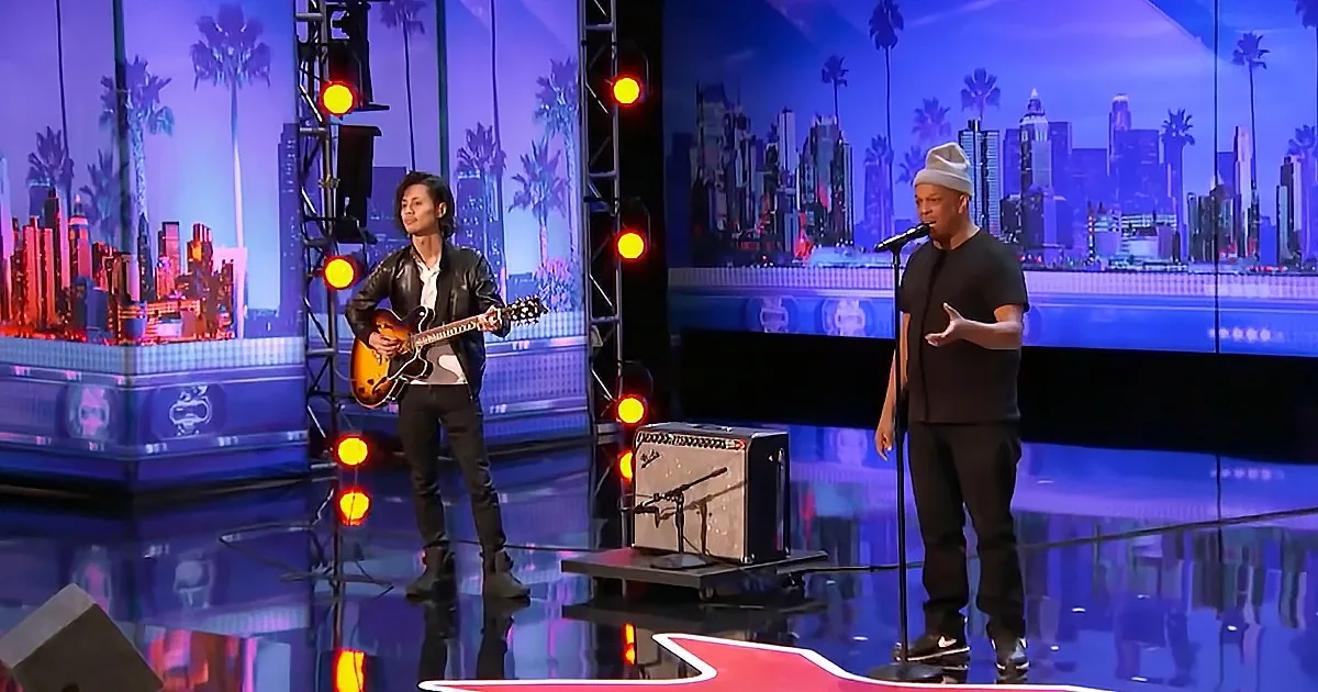 A man in a beanie sings into a microphone while another plays guitar on a stage with a cityscape backdrop.