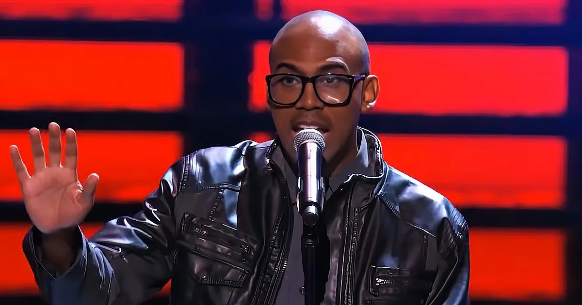 A man in a black leather jacket speaks into a microphone, gesturing with his hand.