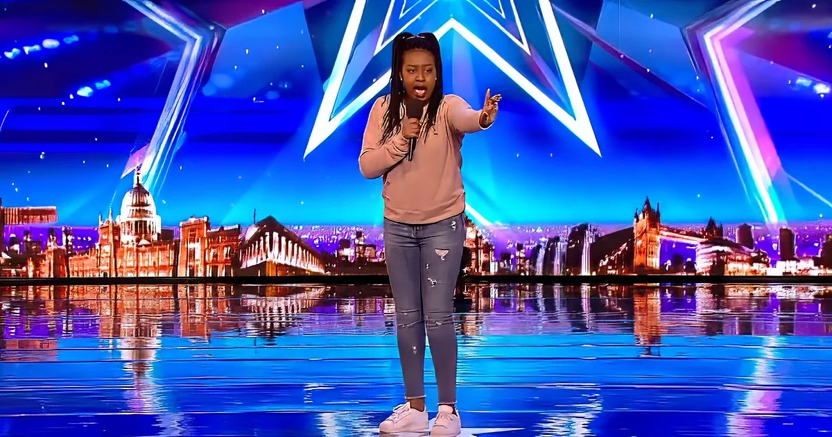 A person stands on a stage with a microphone, gesturing, against a backdrop of a cityscape and blue lighting.