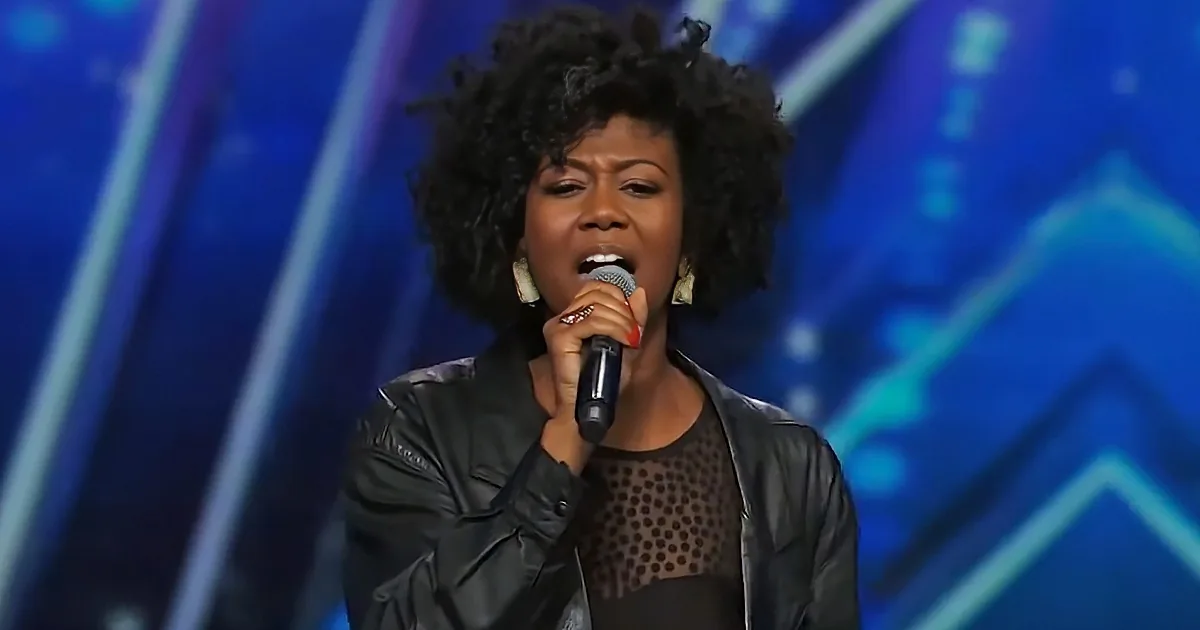 A woman with curly hair sings into a microphone on a stage with blue lighting.
