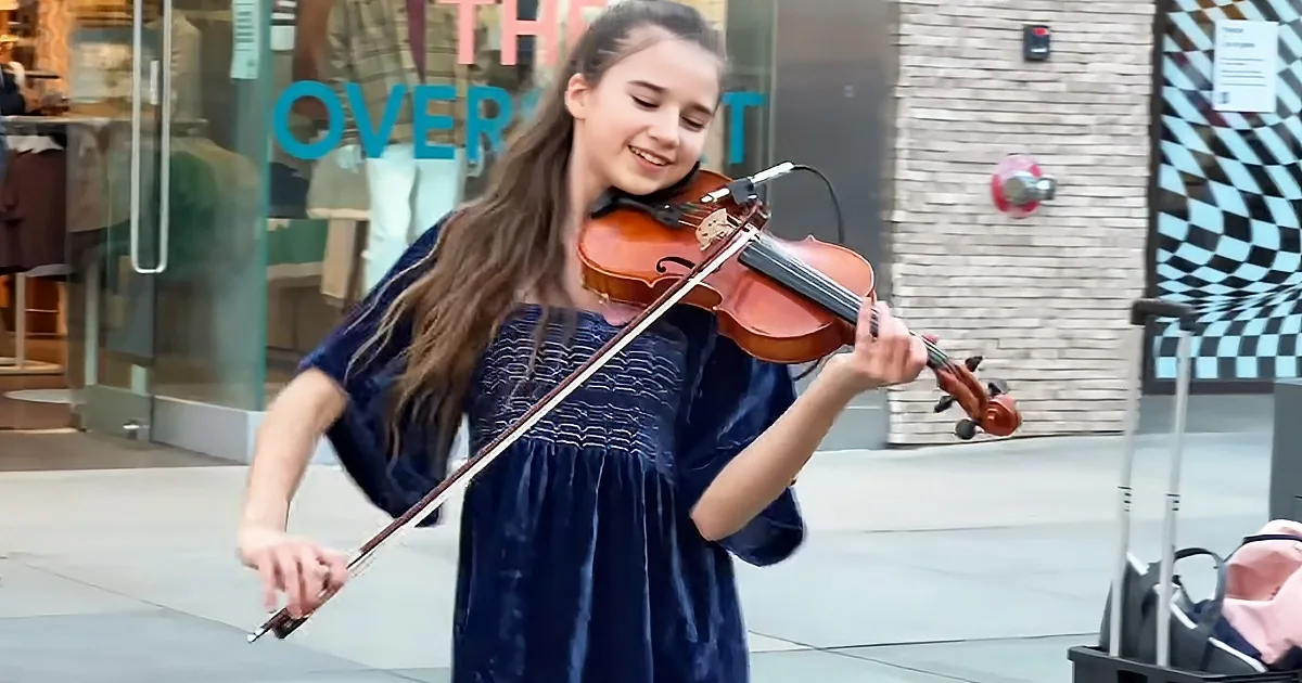A girl in a blue dress plays the violin in front of a store with a checkered window.