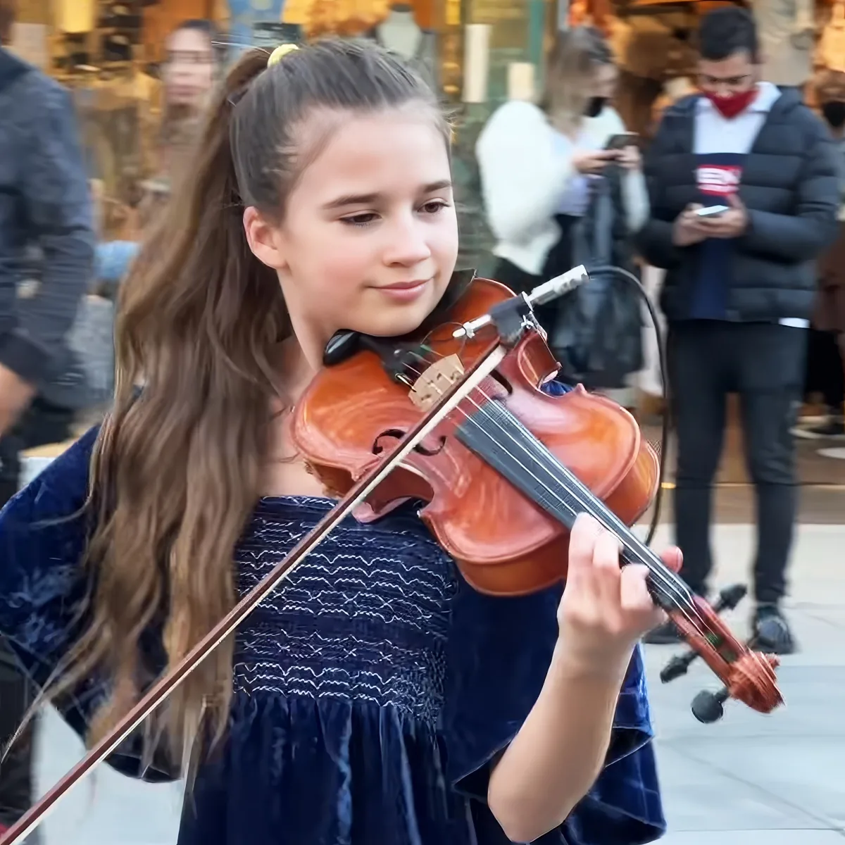 A young girl with long brown hair plays a violin in a public setting, surrounded by people.