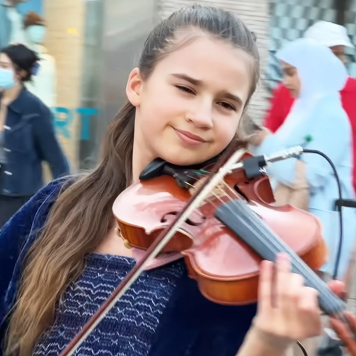 A young girl with long hair smiles while playing a violin outdoors.