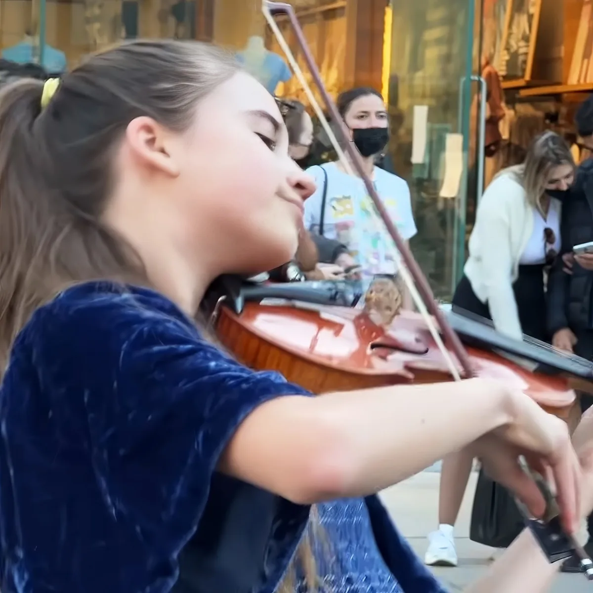 A young woman in a blue shirt plays a violin in a public setting, with people in the background.