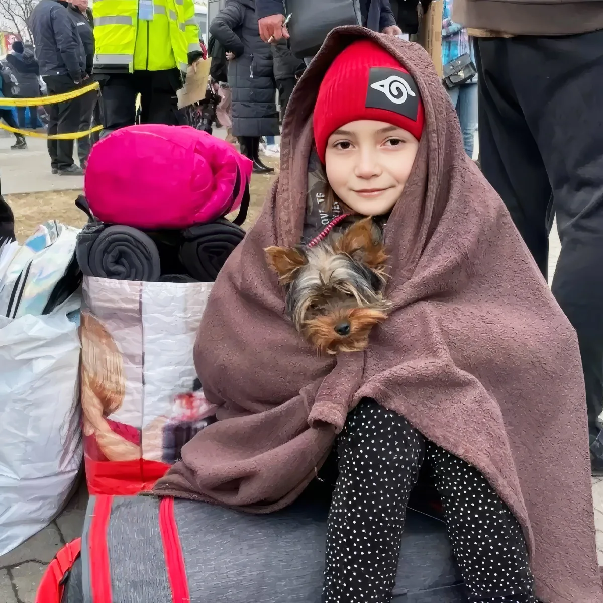 A child in a red beanie and polka-dot pants holds a small dog wrapped in a brown blanket, surrounded by bags and people...