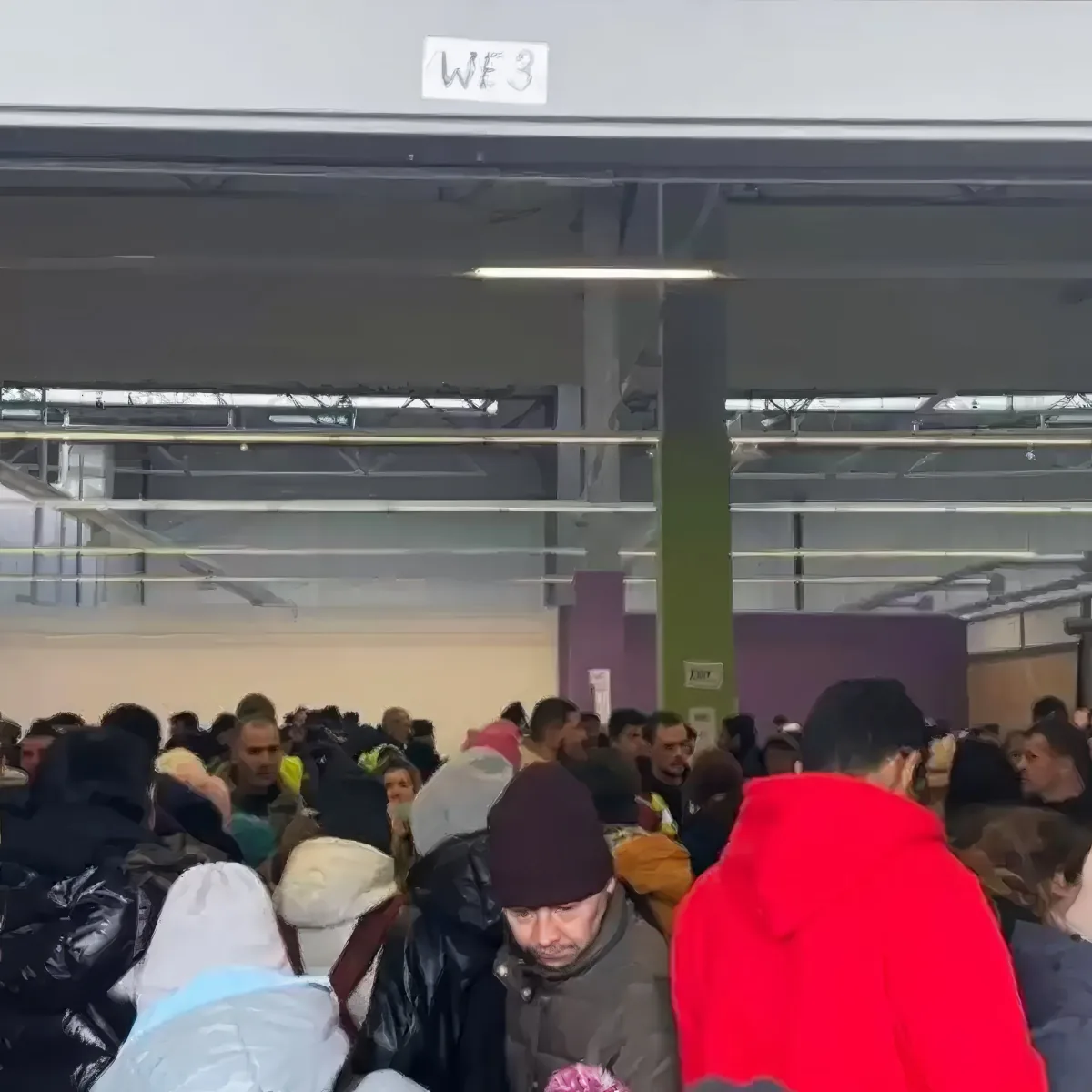 A large crowd of people is gathered in a spacious indoor area with a sign reading "WE3" above.