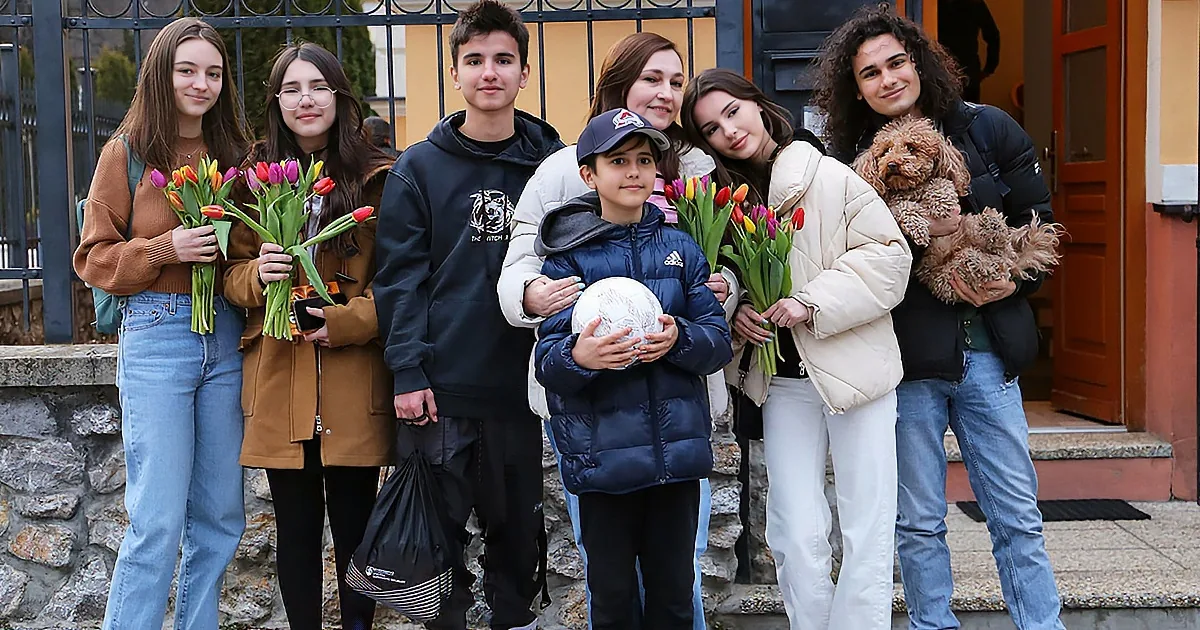A group of seven people, including a child with a soccer ball, stands outside holding flowers and a dog, smiling.