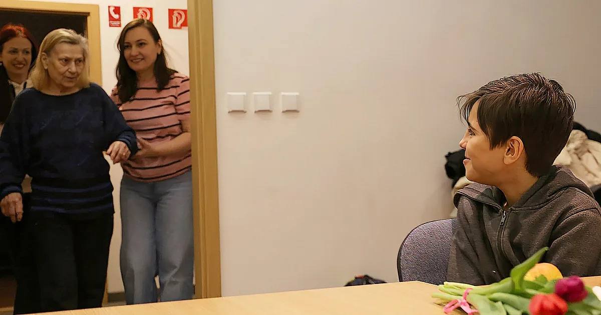 A person in a grey hoodie sits at a table, while two women stand in a doorway, smiling.