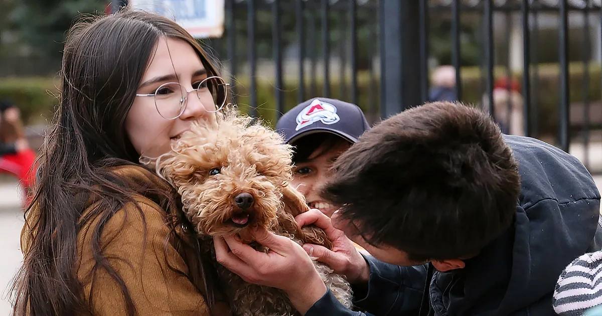 A woman holds a fluffy dog while another person leans in close, both outdoors near a fence.