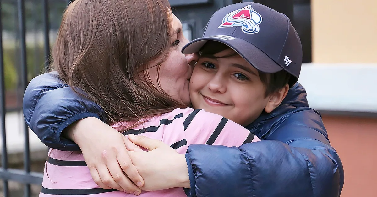 A woman hugs a young boy wearing a Colorado Avalanche cap, both dressed warmly, with a fence and building in the background.