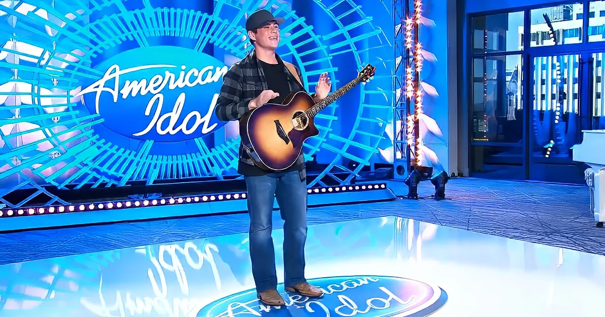 A man in a plaid shirt and jeans holds a guitar on stage at American Idol.