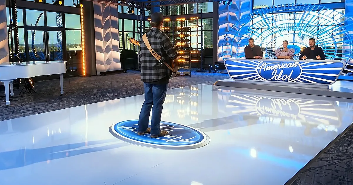 A man in a plaid shirt and jeans stands on a stage with a guitar, facing judges on a set resembling "American Idol."