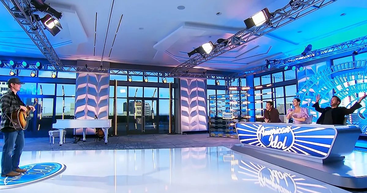 A man plays guitar on stage at American Idol, judges react, blue lighting, cityscape view.