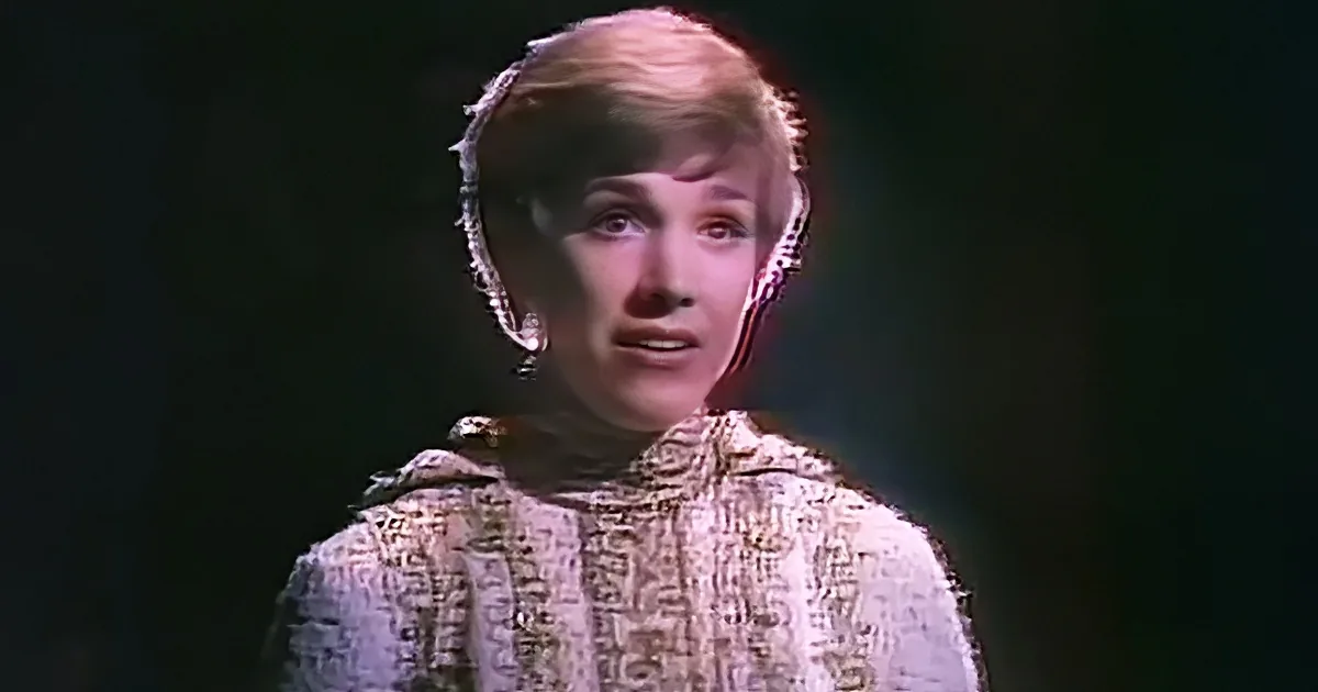 A person wearing a patterned outfit and a head covering is shown against a dark background.