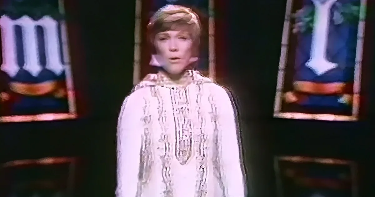 A person in a white, patterned outfit stands in front of stained glass windows with a dark background.