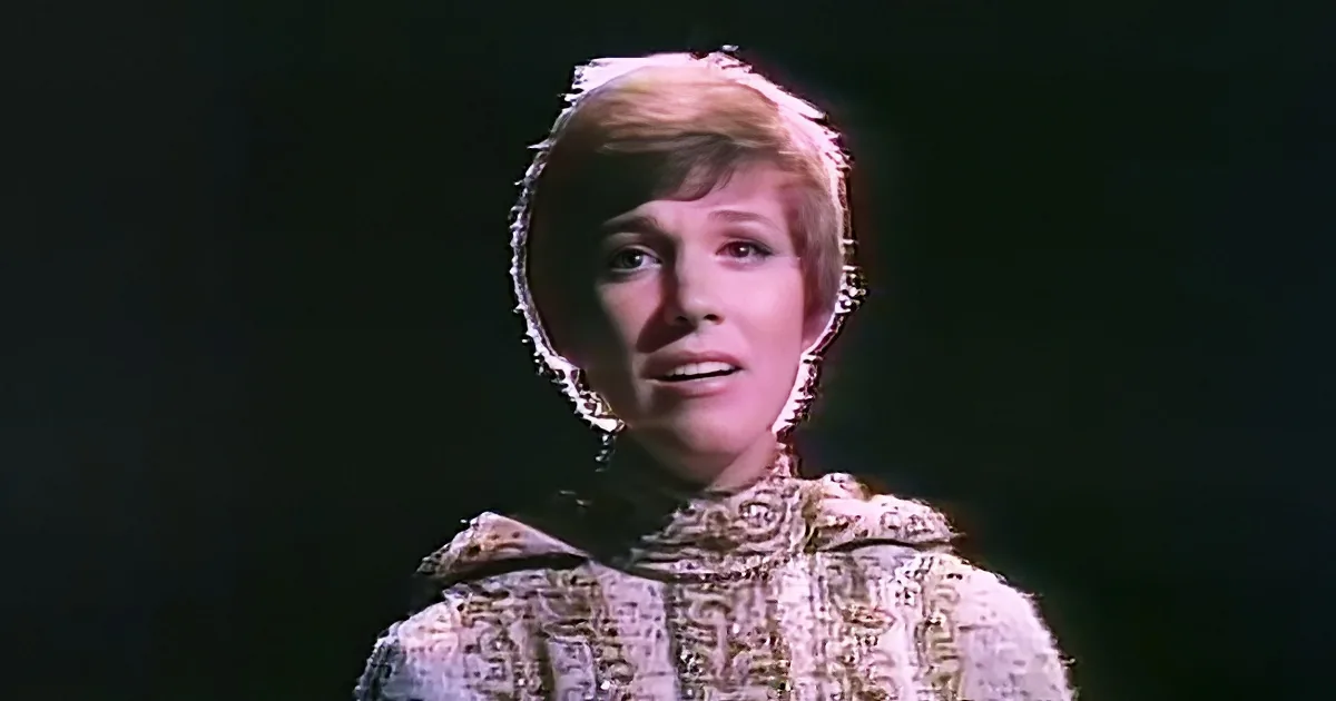 A woman with short, styled hair and a patterned outfit is seen against a dark background.