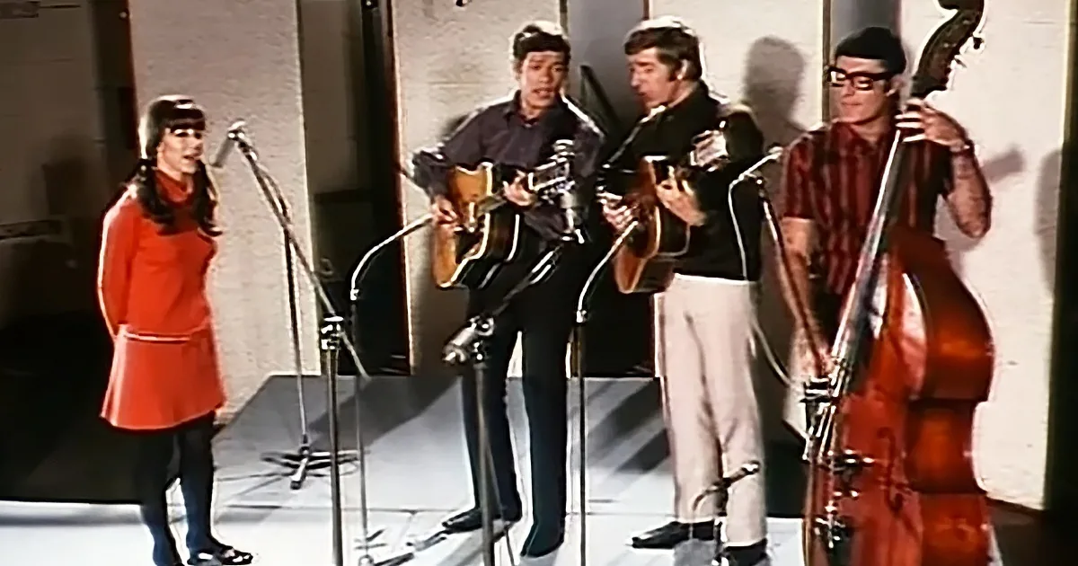 A band performs on stage with a woman in a red dress singing, two guitarists, and a bassist.