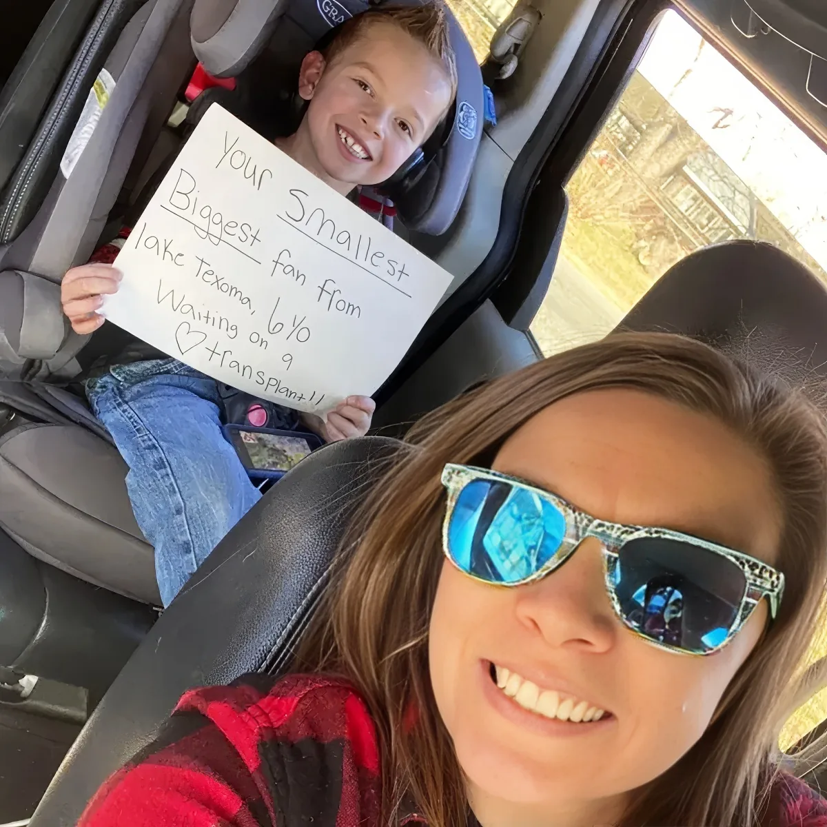 A woman in sunglasses smiles while a child in a car seat holds a sign about a transplant.