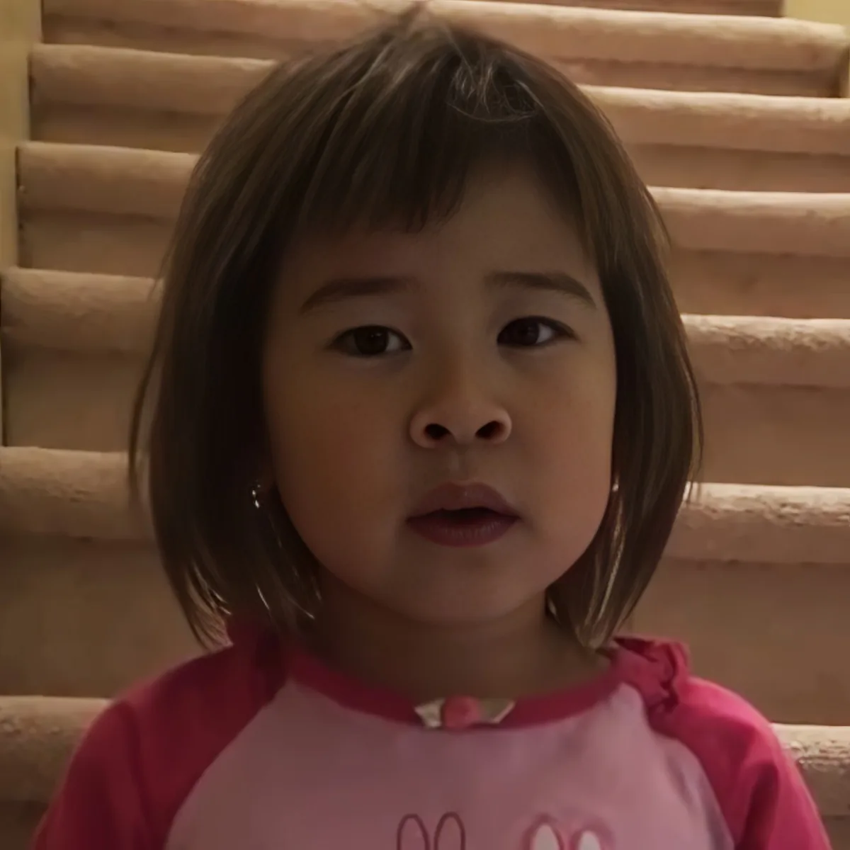 A young child with short brown hair, wearing a pink shirt, sits in front of a staircase.