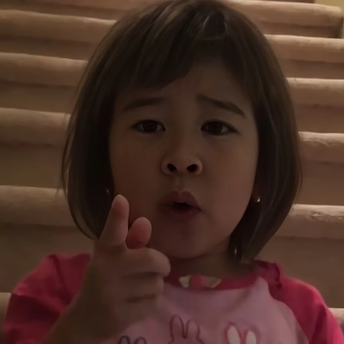 A young child with short hair, wearing a pink shirt, gives a thumbs-up gesture.