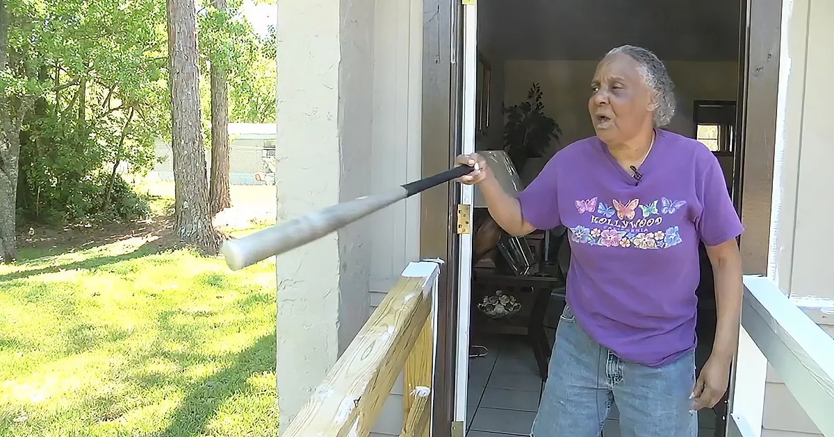 A person in a purple shirt stands on a porch, holding a baseball bat, with a grassy yard and trees in the background.