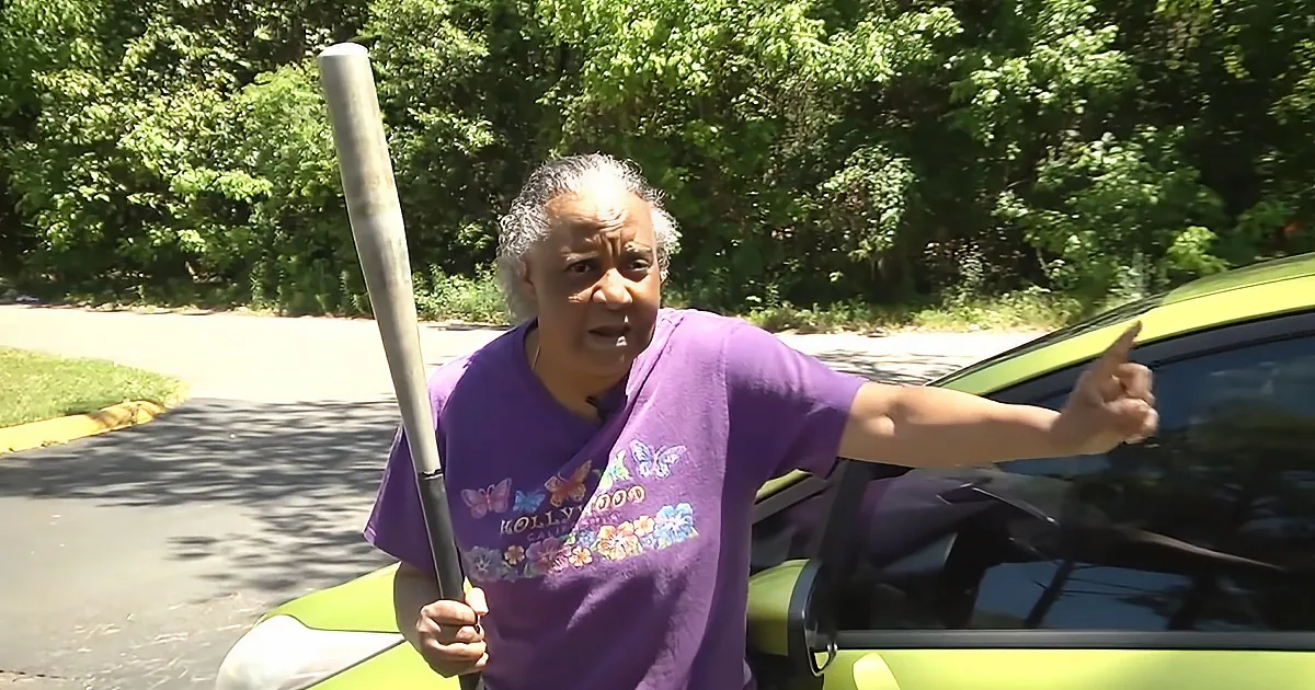 A woman in a purple shirt holds a baseball bat and points while standing near a green car.