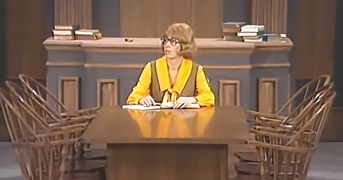 A person in a yellow robe sits at a desk with a book, surrounded by chairs and a backdrop resembling a courtroom.