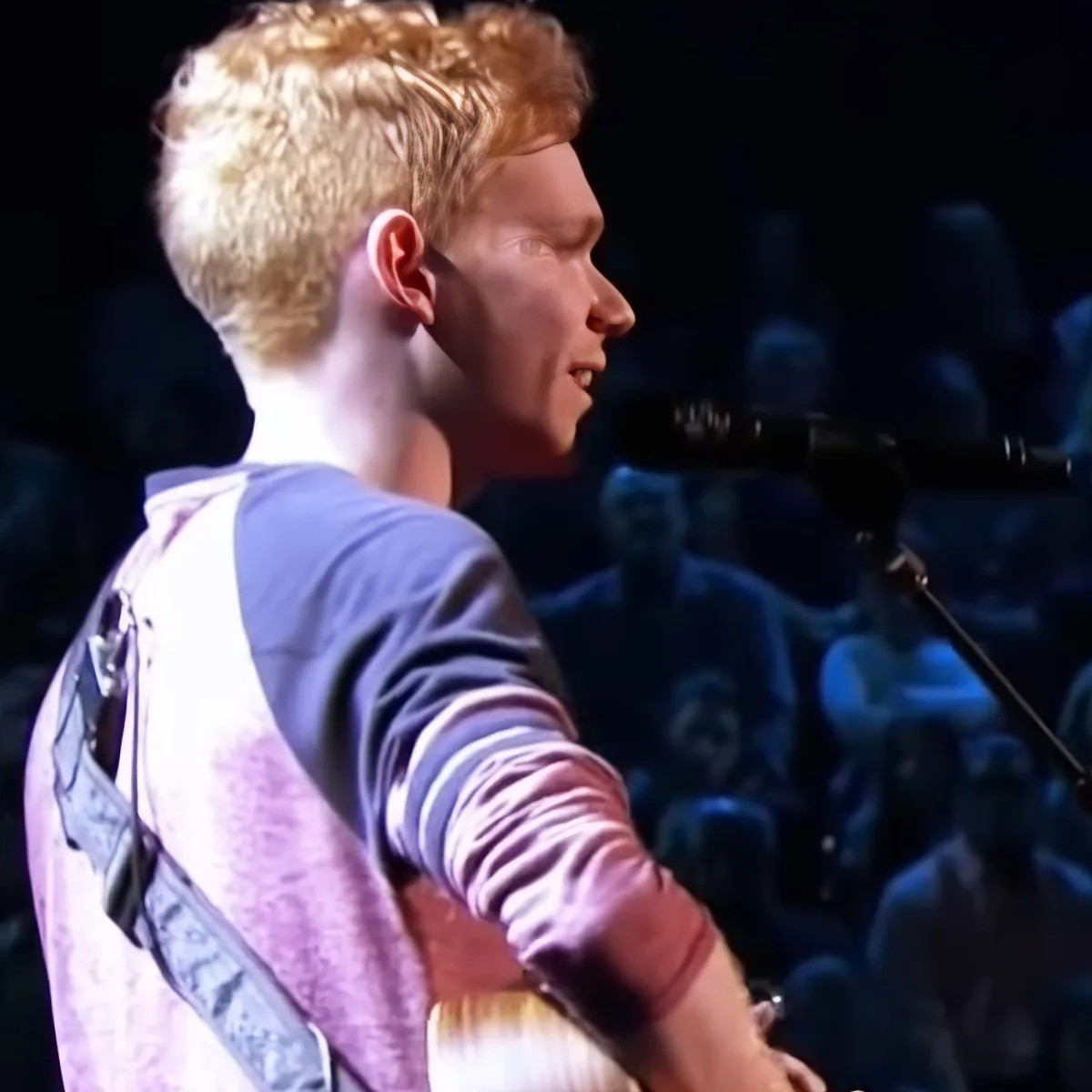 A person with blonde hair, wearing a striped shirt, is singing into a microphone while holding a guitar.