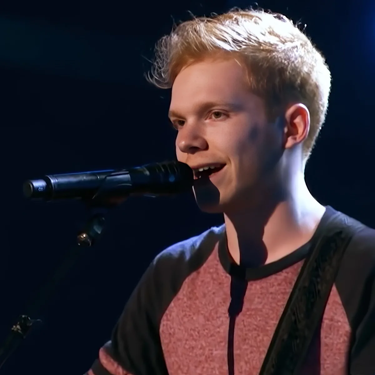 A young person with short, light-colored hair sings into a microphone, wearing a red and black shirt.
