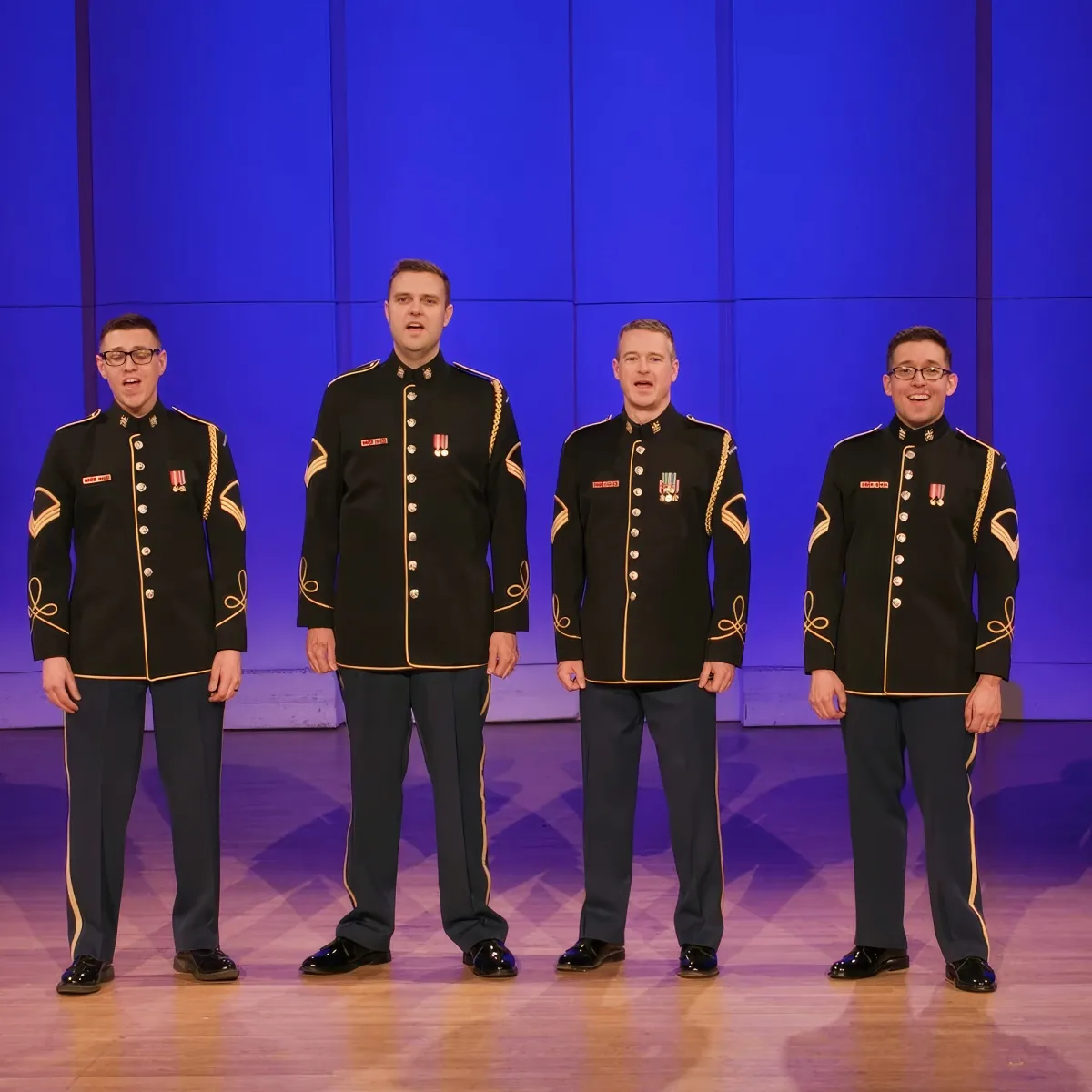 Four men in black military uniforms with gold trim stand in a row on a stage.