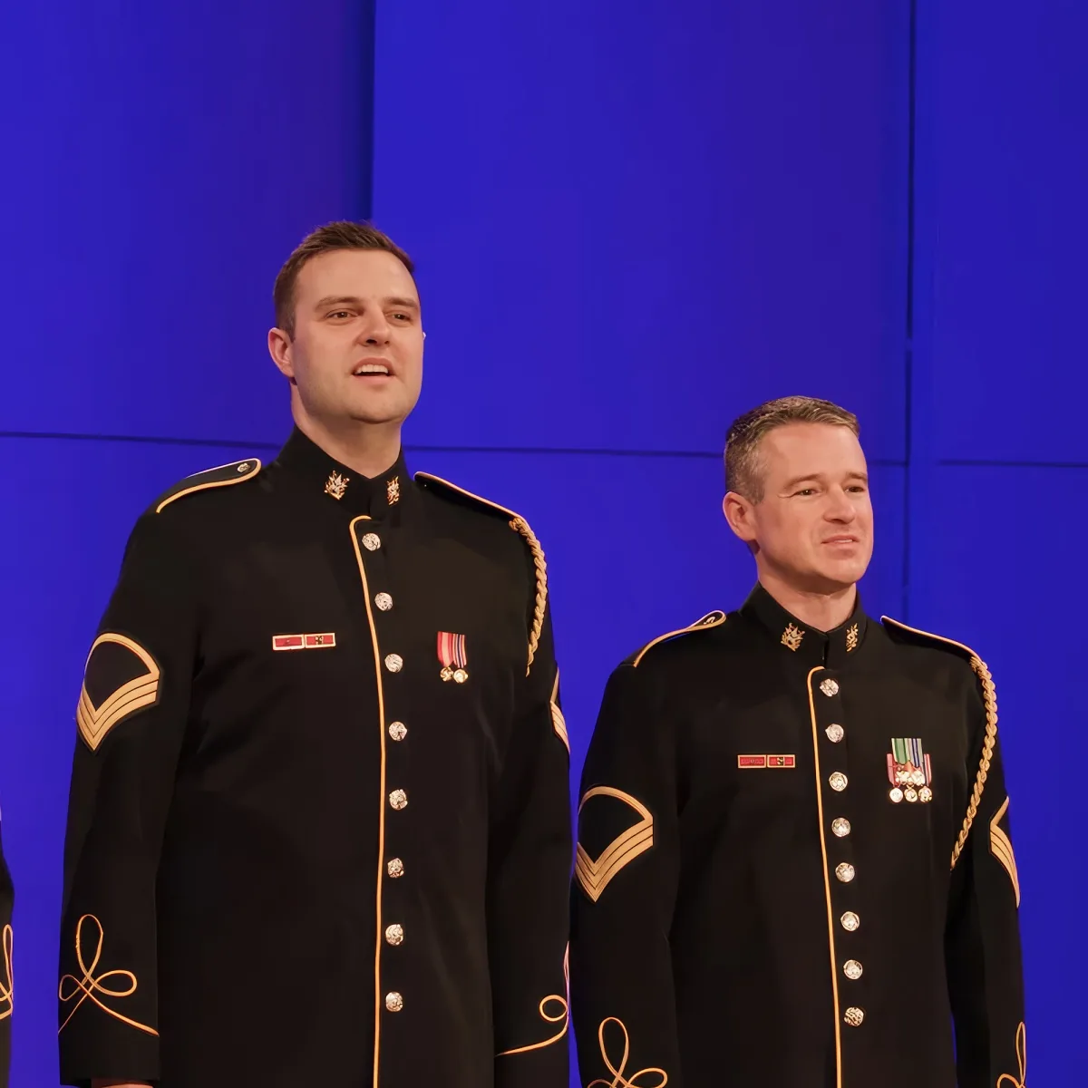 Two men in black military uniforms with gold trim and medals stand side by side against a blue background.