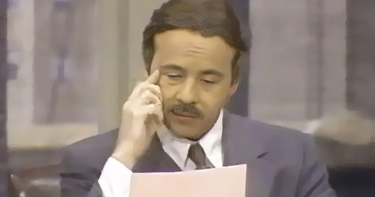 A man in a suit holds a document and gestures with his finger near his temple.