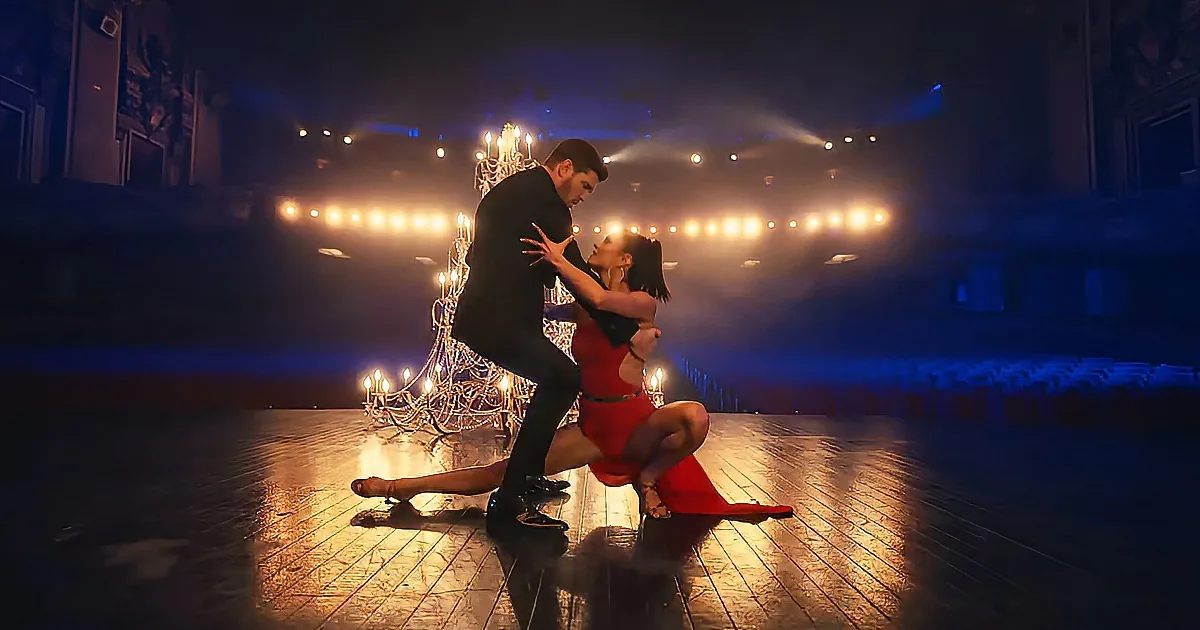 A couple in a dramatic dance pose on a stage with a chandelier and bright lights in the background.