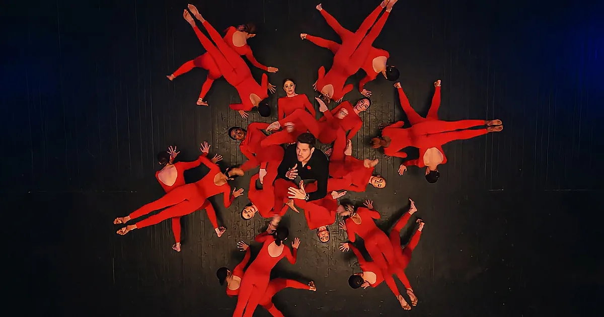 A man in black stands surrounded by dancers in red, forming a star-like pattern on stage.