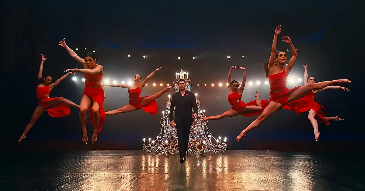 A man in black walks confidently on stage as six dancers in red dresses leap gracefully around him.
