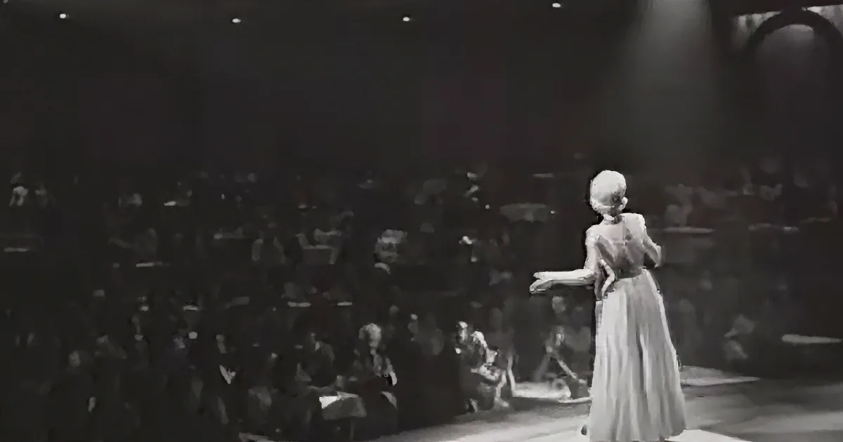 A woman in a long dress stands on stage, gesturing, with an orchestra and audience in the background.