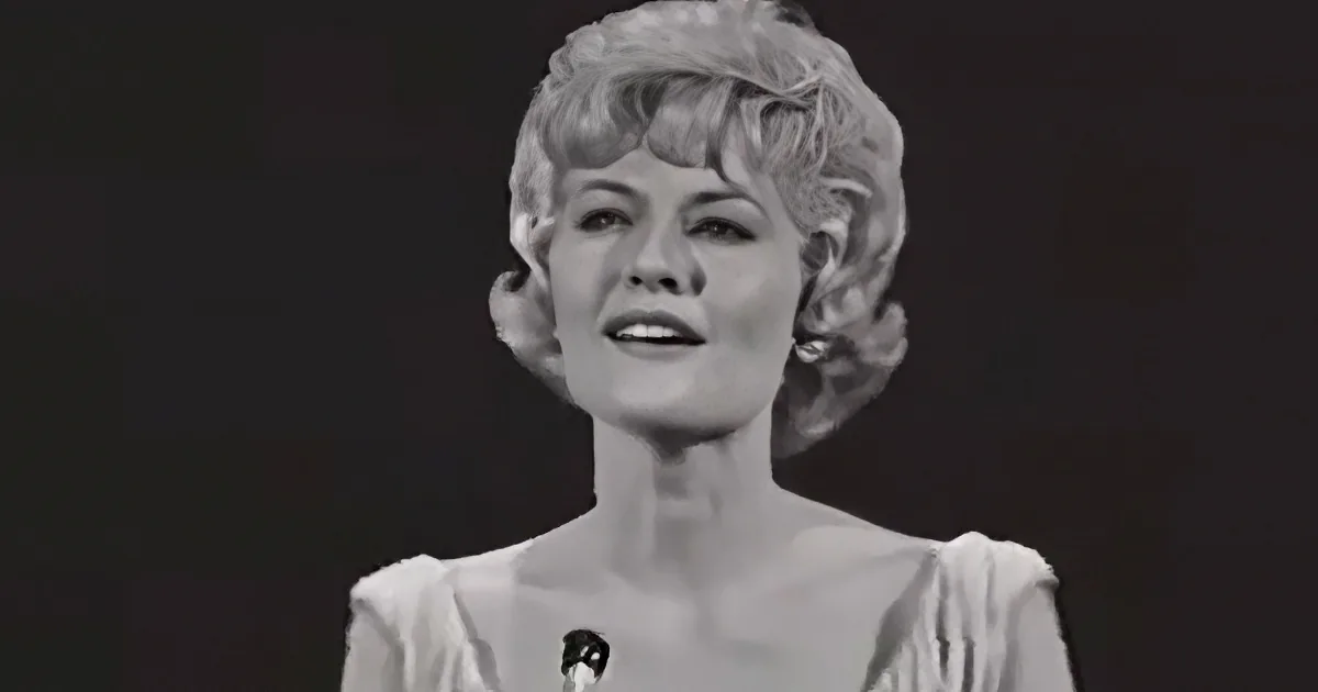 A woman with short, styled hair smiles while speaking into a microphone, wearing a light-colored top.