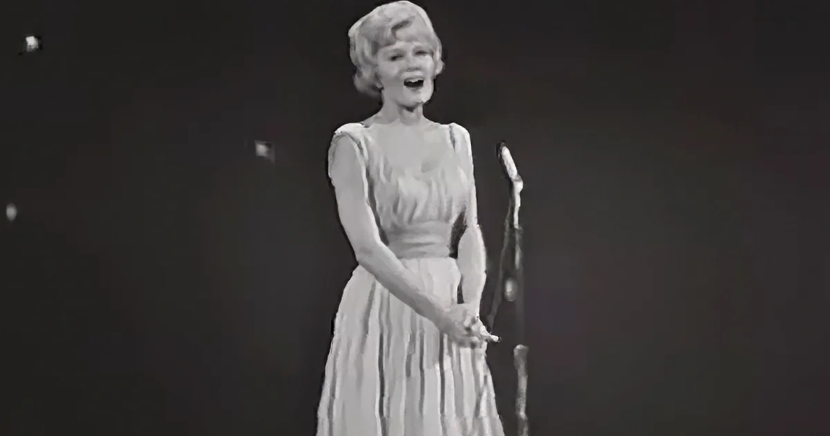 A woman in a white dress sings on stage, holding a microphone.